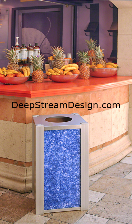 A modern trash receptacle by DeepStream Design with Blue Glass 3form panels in front of a colorful fruit drink stand